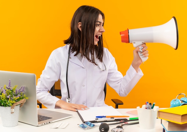 Jeune femme médecin en blouse blanche avec stéthoscope criant au mégaphone avec une expression agressive assis à la table avec un ordinateur portable et des documents sur un mur orange