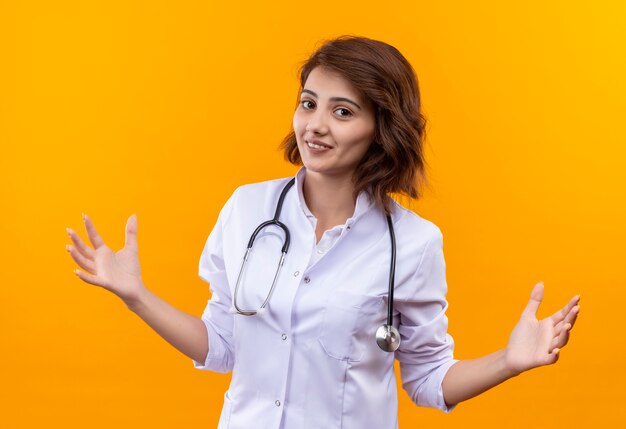 Jeune femme médecin en blouse blanche avec stéthoscope faisant geste de bienvenue étendant les bras souriant debout sur le mur orange