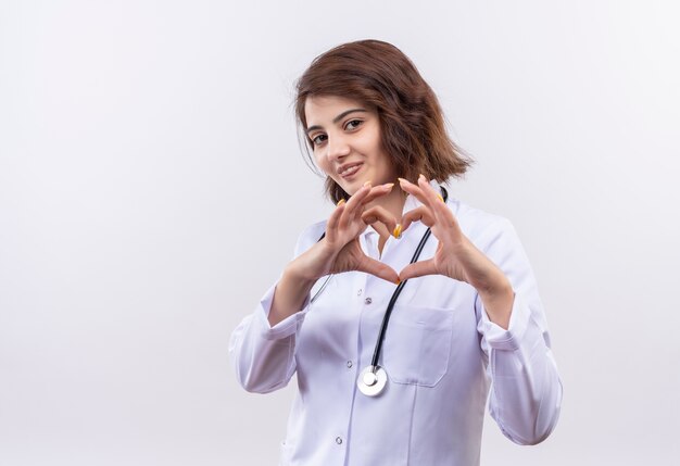 Jeune femme médecin en blouse blanche avec stéthoscope faisant le geste du cœur avec les doigts sur la poitrine souriant debout sur un mur blanc