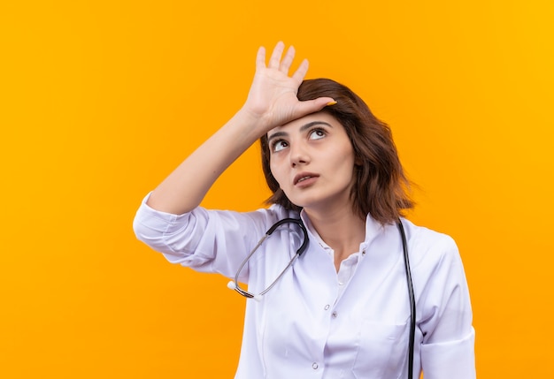 Jeune femme médecin en blouse blanche avec stéthoscope faisant un geste plus lâche avec la main sur sa tête debout sur un mur orange