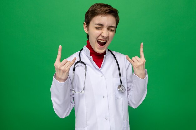 Jeune femme médecin en blouse blanche avec stéthoscope heureux et excité de crier montrant le symbole du rock