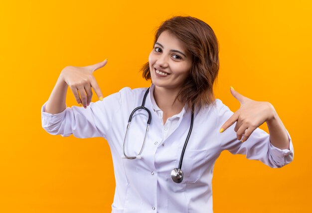 Jeune femme médecin en blouse blanche avec stéthoscope heureux et positif pointant vers elle-même