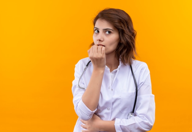 Jeune femme médecin en blouse blanche avec stéthoscope inquiet et nerveux se mordant les ongles debout sur un mur orange