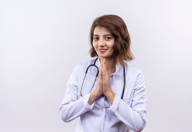 Jeune femme médecin en blouse blanche avec stéthoscope main dans la main dans le geste de prière, se sentir reconnaissant et heureux