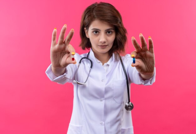 Jeune femme médecin en blouse blanche avec stéthoscope montrant des pilules dans les mains avec un visage sérieux debout sur un mur rose