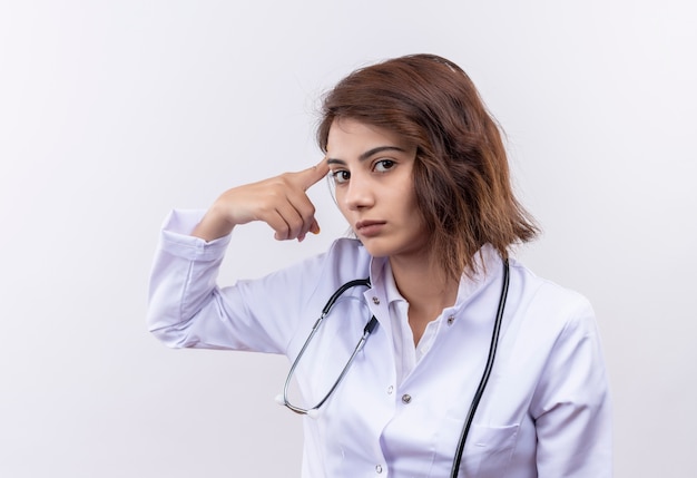 Jeune femme médecin en blouse blanche avec stéthoscope regardant la caméra avec une expression sérieuse en pointant sa tempe