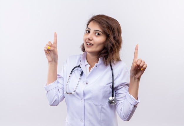 Jeune femme médecin en blouse blanche avec stéthoscope regardant la caméra avec sourire confiant pointant avec finfers up