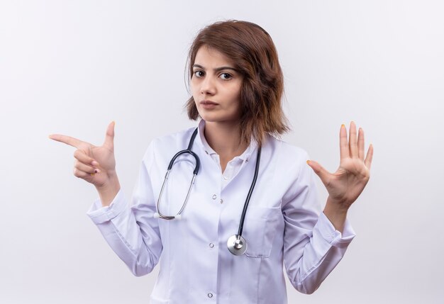 Jeune femme médecin en blouse blanche avec stéthoscope regardant la caméra avec un visage sérieux montrant la main ouverte pointant avec l'index sur le côté