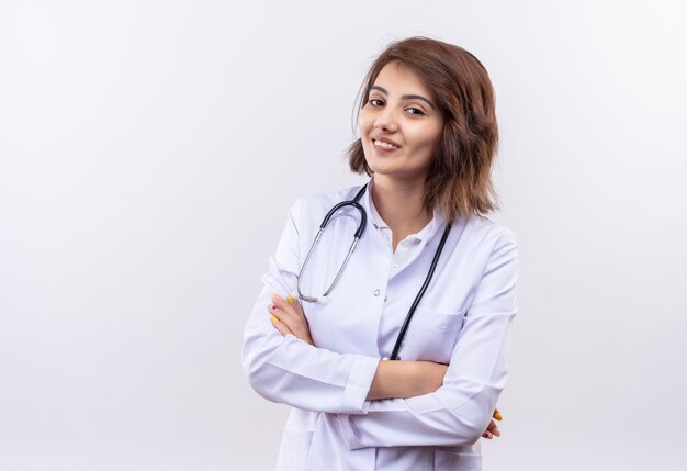 Jeune femme médecin en blouse blanche avec stéthoscope souriant debout confiant avec les bras croisés sur mur blanc