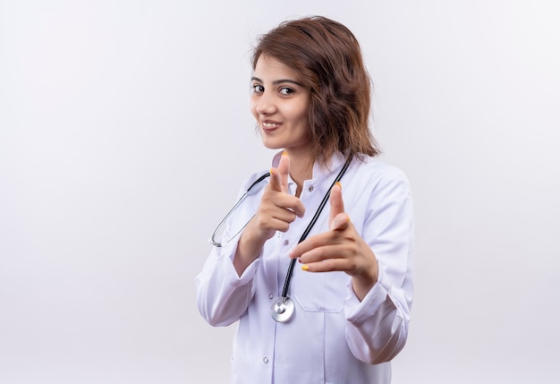 Jeune femme médecin en blouse blanche avec stéthoscope souriant joyeusement pointant avec l'index avec les deux mains debout sur un mur blanc