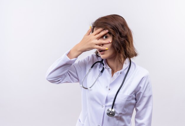 Jeune femme médecin en blouse blanche avec stéthoscope surpris et inquiet couvrant les yeux avec des paumes à la recherche à travers les doigts debout sur un mur blanc