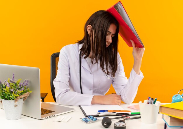 Jeune femme médecin en blouse blanche avec stéthoscope tenant le dossier sur sa tête à la fatigue et surmené assis à la table avec ordinateur portable et documents sur mur orange