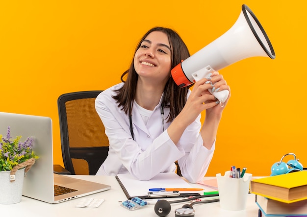 Jeune femme médecin en blouse blanche avec stéthoscope tenant un mégaphone souriant avec un visage heureux assis à la table avec un ordinateur portable et des documents sur un mur orange