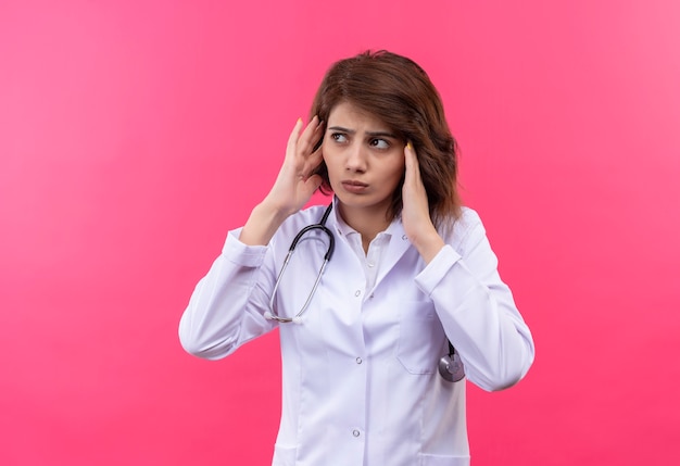 Jeune femme médecin en blouse blanche avec stéthoscope touchant les tempes avec les mains à la fatigue ayant mal de tête debout sur le mur rose