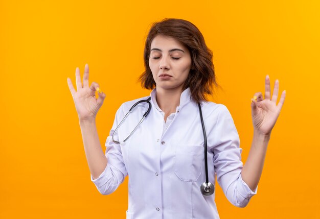 Jeune femme médecin en blouse blanche avec stéthoscope avec les yeux fermés relaxant faisant le geste de méditation avec les doigts debout sur le mur orange