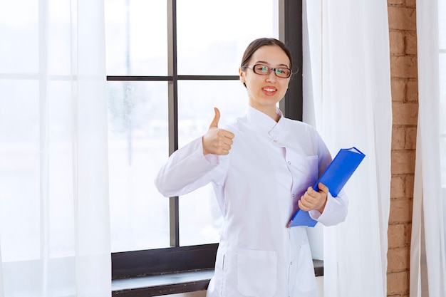 Photo gratuite jeune femme médecin en blouse blanche tenant un classeur dans ses mains près de la fenêtre.