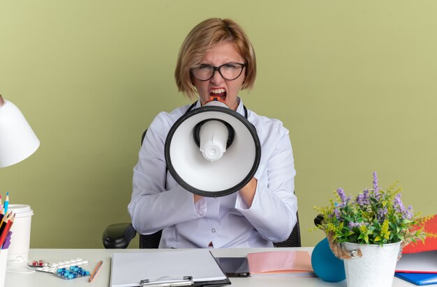 Une jeune femme médecin en colère portant une robe médicale avec des lunettes et un stéthoscope est assise à table avec des outils médicaux parle sur un haut-parleur isolé sur un mur vert olive