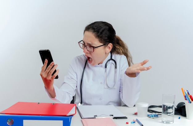 Jeune femme médecin en colère portant une robe médicale et un stéthoscope et des lunettes assis au bureau avec des outils médicaux tenant et regardant un téléphone mobile montrant une main vide isolée