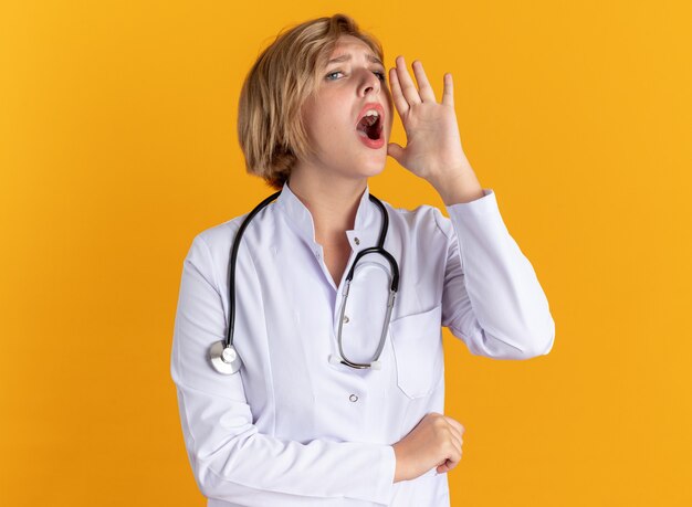 Jeune femme médecin concernée portant une robe médicale avec stéthoscope appelant quelqu'un isolé sur un mur orange