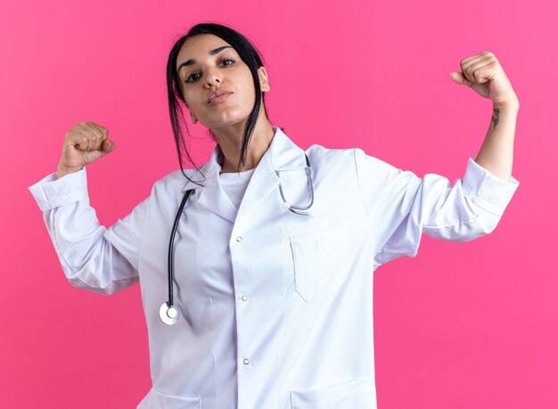 Jeune femme médecin confiante portant une robe médicale avec stéthoscope montrant un geste fort isolé sur un mur rose