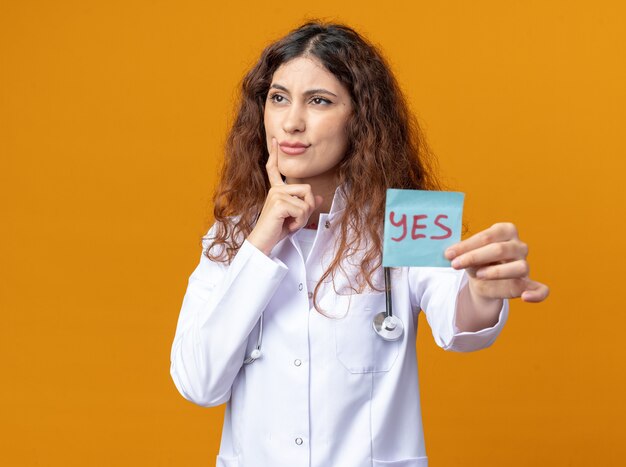 Jeune femme médecin confuse portant une robe médicale et un stéthoscope regardant le côté s'étirant oui note en gardant le doigt sur le visage isolé sur un mur orange avec espace de copie