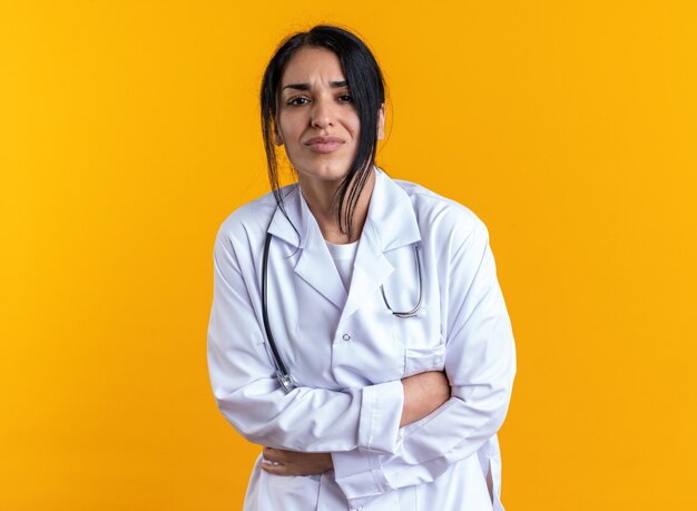 Une jeune femme médecin douloureuse portant une robe médicale avec un stéthoscope saisi l'estomac isolé sur un mur jaune