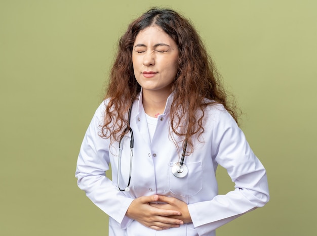 Jeune Femme Médecin Douloureuse Portant Une Robe Médicale Et Un Stéthoscope Tenant Le Ventre Avec Les Yeux Fermés Isolés Sur Un Mur Vert Olive