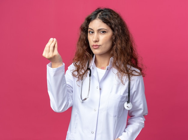 Une jeune femme médecin douteuse portant une robe médicale et un stéthoscope aux lèvres pincées fait semblant de tenir quelque chose d'isolé sur un mur rose