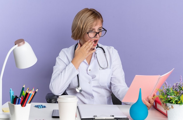 Une jeune femme médecin effrayée portant une robe médicale avec un stéthoscope et des lunettes est assise à table avec des outils médicaux tenant et regardant un ordinateur portable isolé sur fond bleu