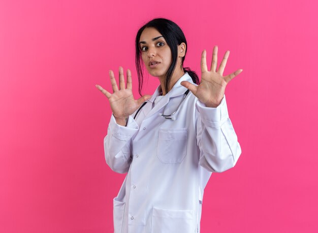 Jeune femme médecin effrayée portant une robe médicale avec stéthoscope montrant un geste d'arrêt isolé sur fond rose