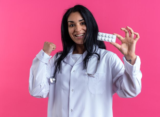 Jeune femme médecin enthousiaste portant une robe médicale avec un stéthoscope tenant des pilules montrant un geste oui isolé sur un mur rose