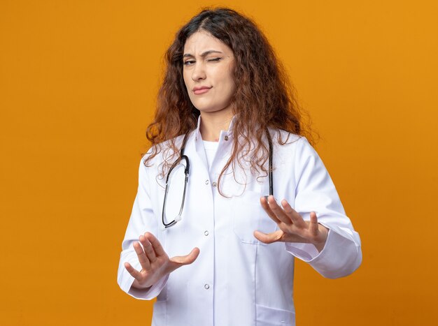 Jeune femme médecin fronçant les sourcils portant une robe médicale et un stéthoscope faisant un geste de refus avec un œil fermé isolé sur un mur orange avec espace de copie