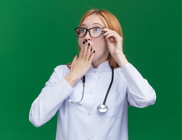 Jeune femme médecin gingembre concerné portant une robe médicale et un stéthoscope avec des lunettes saisissant des lunettes regardant de côté en gardant la main sur la bouche