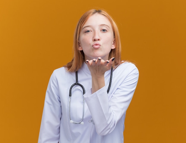 Jeune femme médecin gingembre confiant portant une robe médicale et un stéthoscope envoyant un baiser à la caméra