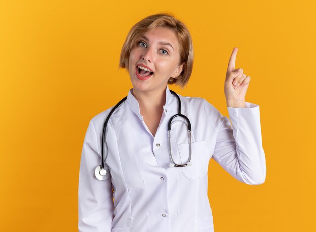 Jeune femme médecin impressionnée portant une robe médicale avec des points de stéthoscope vers le haut isolé sur un mur orange