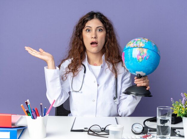 Jeune femme médecin impressionnée portant une robe médicale et un stéthoscope assis à table avec des outils médicaux tenant un globe regardant à l'avant montrant une main vide isolée sur un mur violet