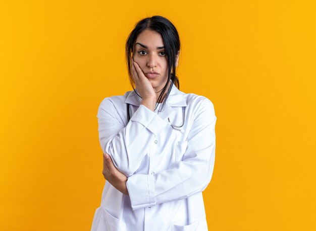 Jeune femme médecin impressionnée portant une robe médicale avec stéthoscope mettant la main sur la joue isolée sur un mur jaune