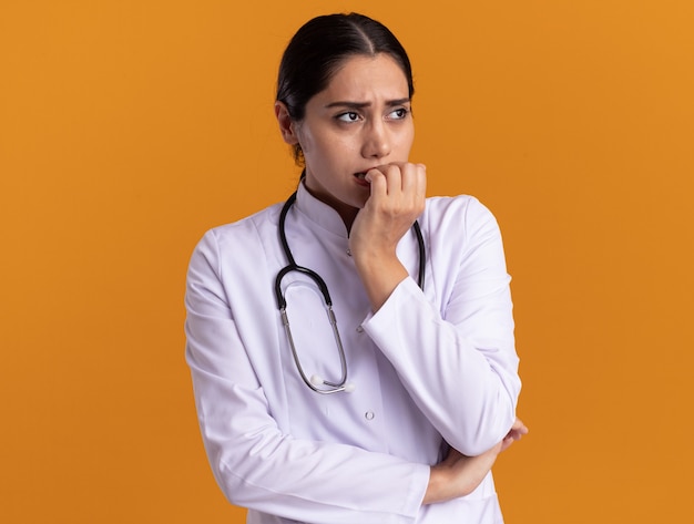 Jeune femme médecin en manteau médical avec stéthoscope autour du cou à côté stressé et nerveux mordant les ongles debout sur un mur orange