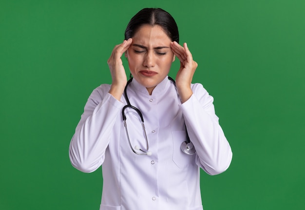 Jeune femme médecin en manteau médical avec stéthoscope autour du cou à la recherche de mal à toucher ses tempes souffrant de maux de tête debout sur un mur vert