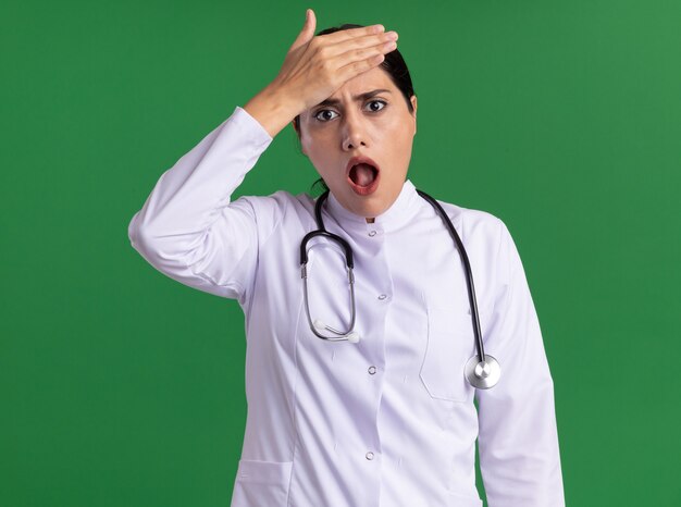 Jeune femme médecin en manteau médical avec stéthoscope à l'avant d'être choqué avec la main sur son front debout sur mur vert
