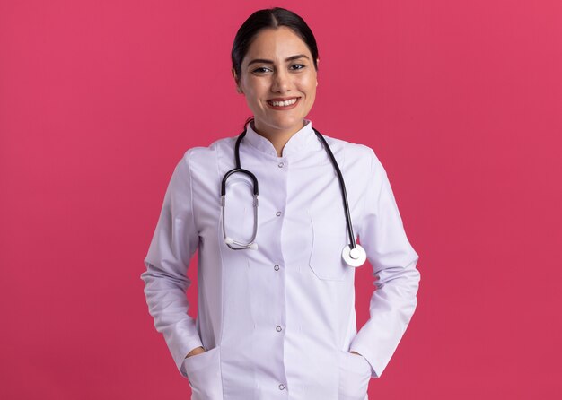 Jeune femme médecin en manteau médical avec stéthoscope à l'avant souriant avec visage heureux debout sur un mur rose