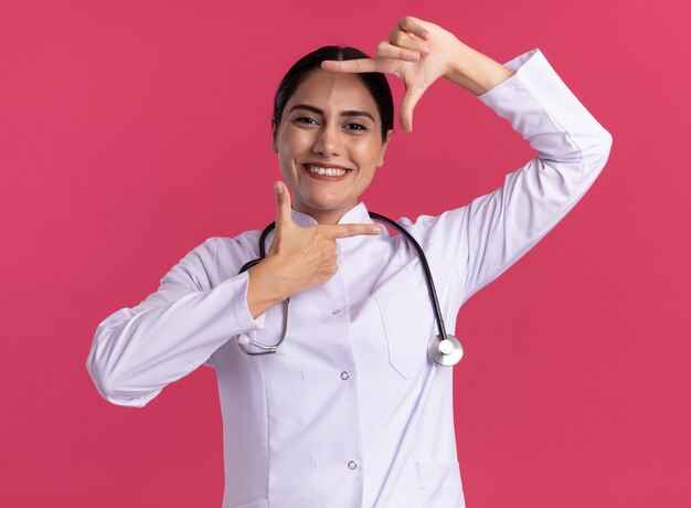 Jeune femme médecin en manteau médical avec stéthoscope à l'avant avec le sourire sur le visage montrant le cadre avec les doigts debout sur le mur rose