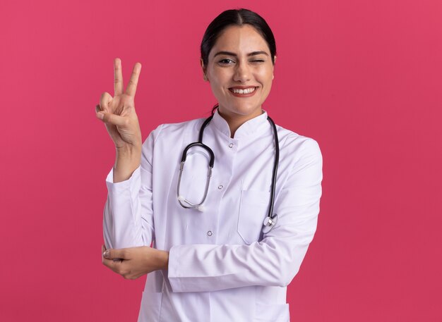 Jeune femme médecin en manteau médical avec stéthoscope à l'avant avec sourire sur le visage montrant v-sign debout sur un mur rose