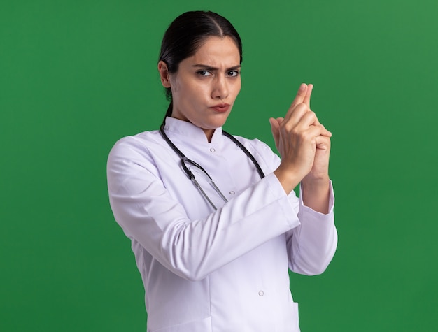 Jeune femme médecin en manteau médical avec stéthoscope à l'avant avec un visage sérieux faisant le geste du pistolet avec les doigts debout sur le mur vert