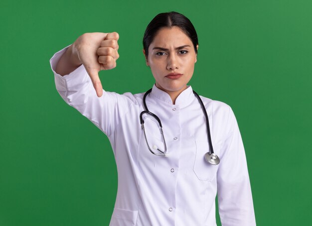 Jeune femme médecin en manteau médical avec stéthoscope à l'avant avec un visage sérieux montrant les pouces vers le bas debout sur un mur vert