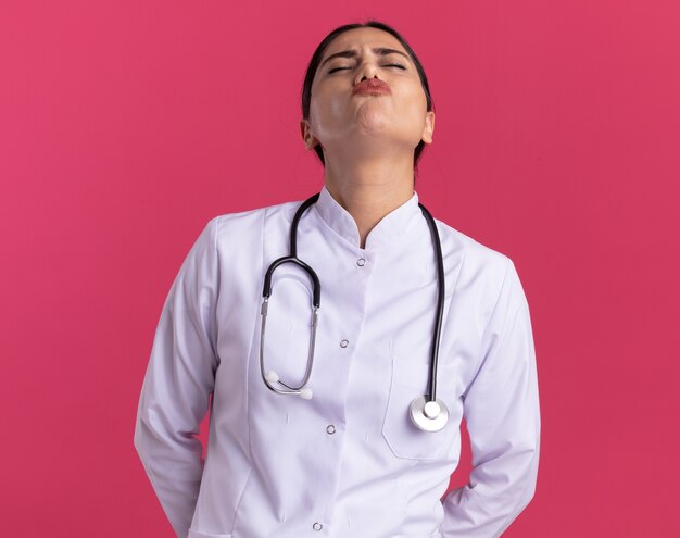 Jeune femme médecin en manteau médical avec stéthoscope avec les yeux fermés avec une expression agacée debout sur un mur rose