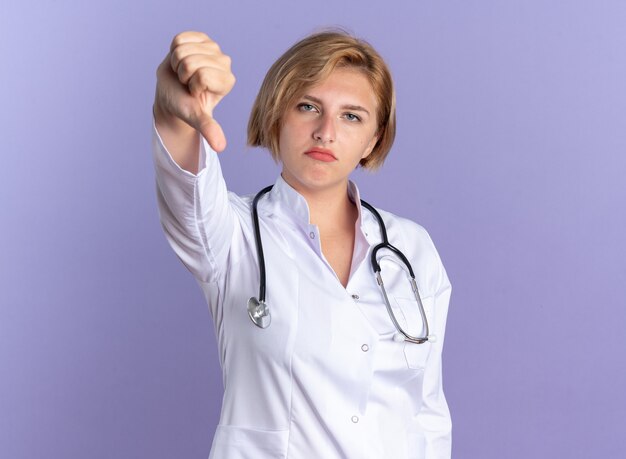 Jeune femme médecin mécontent portant une robe médicale avec stéthoscope montrant le pouce vers le bas isolé sur fond bleu
