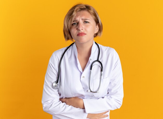 Une jeune femme médecin mécontente portant une robe médicale avec un stéthoscope a attrapé un estomac douloureux isolé sur fond orange