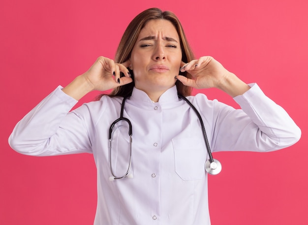 Jeune femme médecin mécontente portant une robe médicale avec stéthoscope oreilles fermées isolées sur mur rose