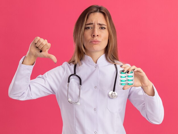 Jeune femme médecin mécontente portant une robe médicale avec stéthoscope tenant des pilules montrant le pouce vers le bas isolé sur un mur rose